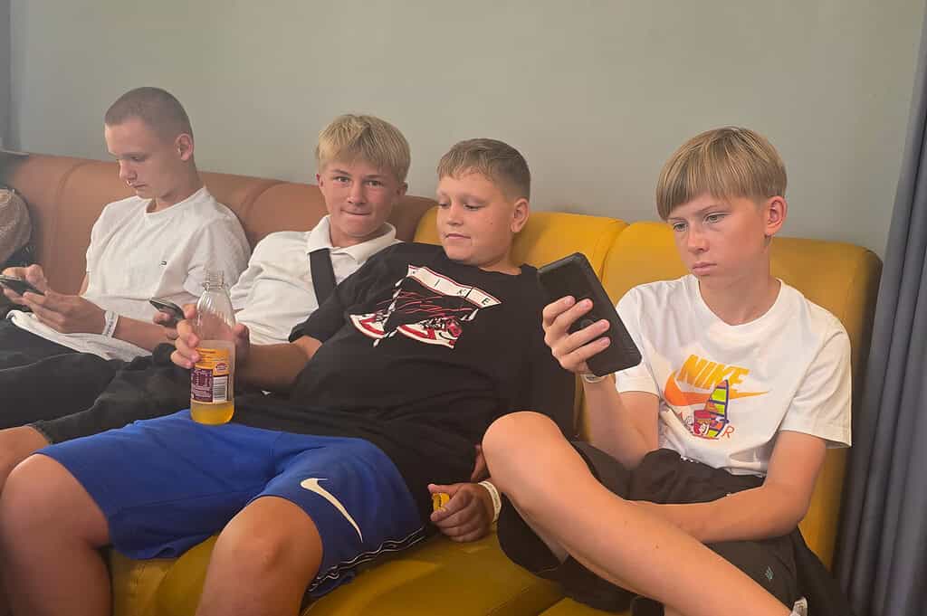 Vier Jugendliche sitzen entspannt auf einem gelben Sofa, schauen auf ihre Smartphones und halten ein Getränk. Die Gesichter wirken ruhig und leicht müde, was die Atmosphäre am Morgen der Heimreise wiedergibt.