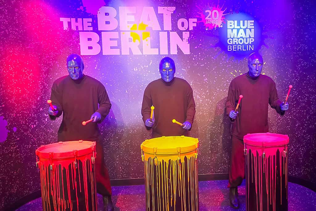 Drei Musiker mit blauer Gesichtsfarbe spielen auf bunten Trommeln bei einem Event in Berlin.