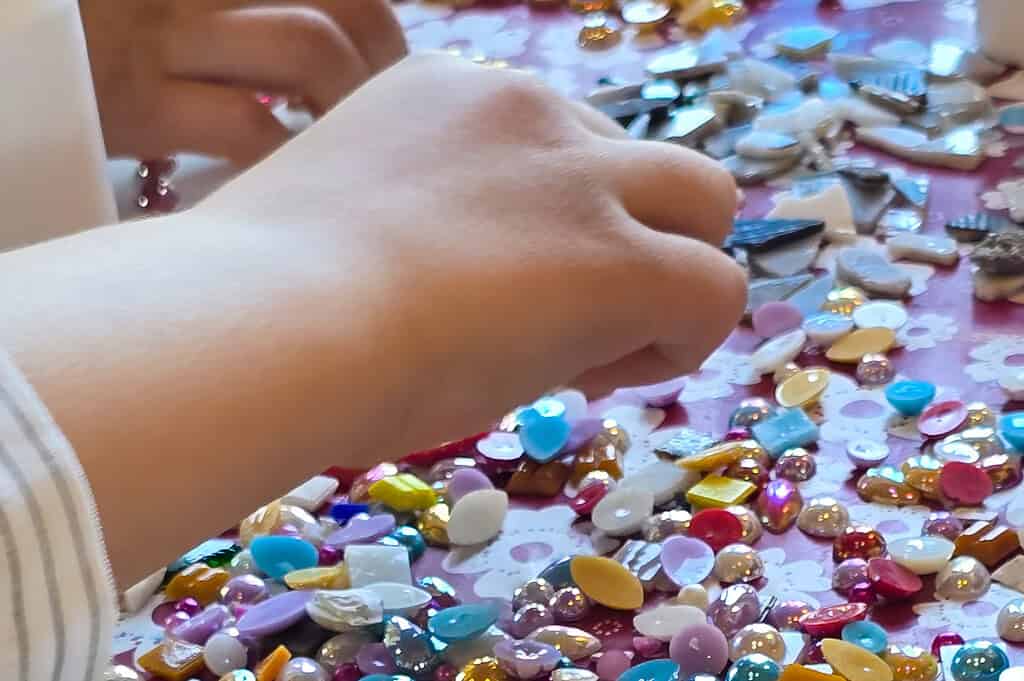 Hände von Kindern sortieren viele kleine, bunte Schmucksteine und Perlen beim Osterferienprojekt Made in Langendorf.